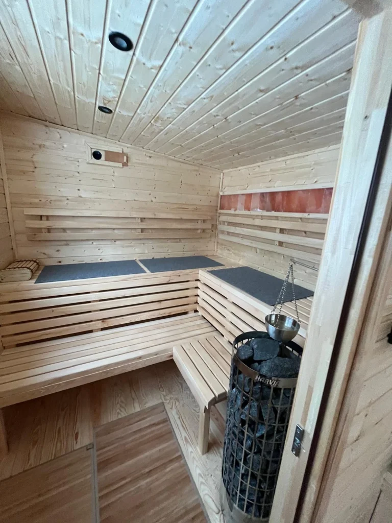 Drewniana ceberka z chochlą do wody stojąca na górnej ławie w saunie, w tle delikatna para unosząca się znad pieca