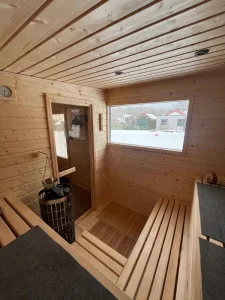 Przytulna, oświetlona ciepłym światłem sauna ogrodowa wieczorową porą, widoczna przez przeszklone drzwi na tle zaśnieżonego ogrodu