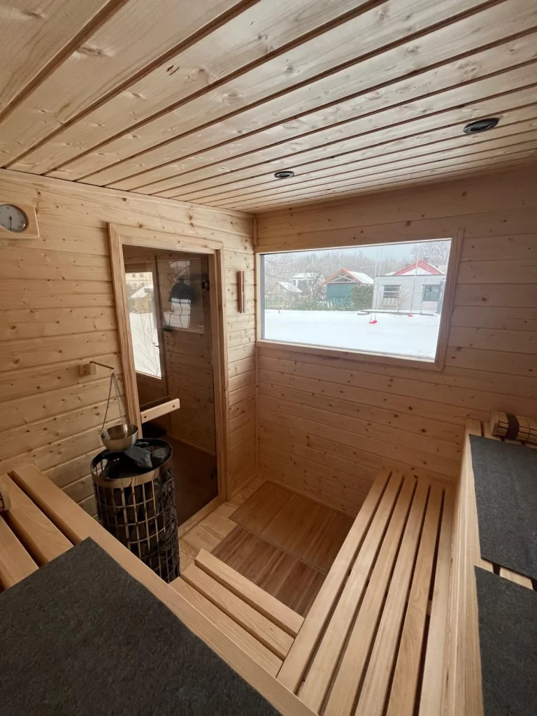 Przytulna, oświetlona ciepłym światłem sauna ogrodowa wieczorową porą, widoczna przez przeszklone drzwi na tle zaśnieżonego ogrodu